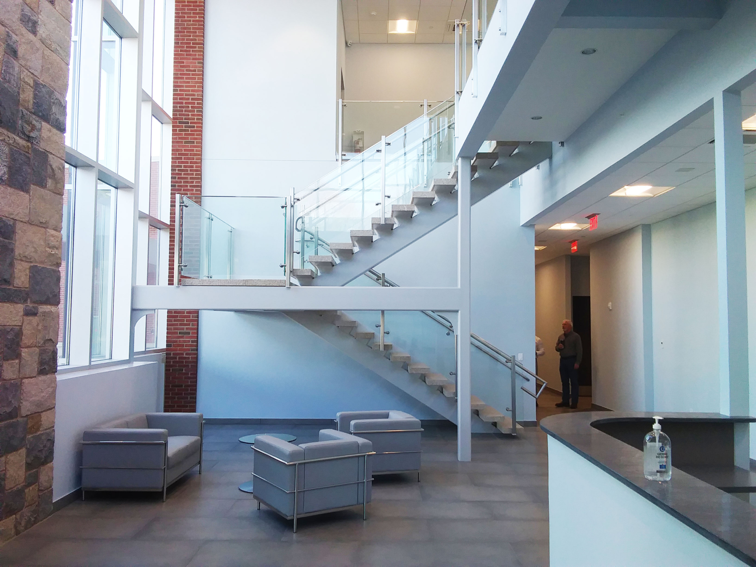 A two-story open office lobby, with white walls, a stone floor and brick trim. An open staircase with clear glass railing panels.