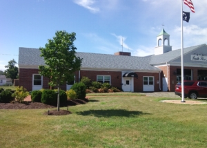 A one-story brick firehouse with bays on the left for firetrucks. An American flag on a flagpole. On the left, a small maple tree and shrubs.